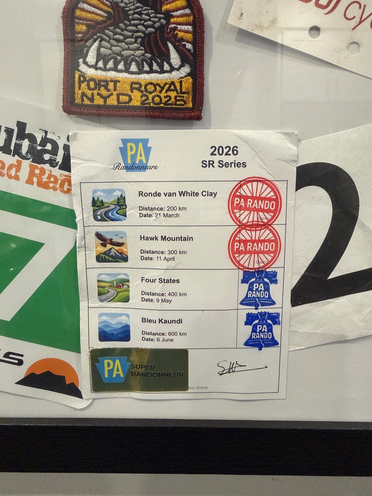 A fully stamped 2026 PA Randonneurs SR Series card, pinned to a board alongside old race numbers and a brevet patch