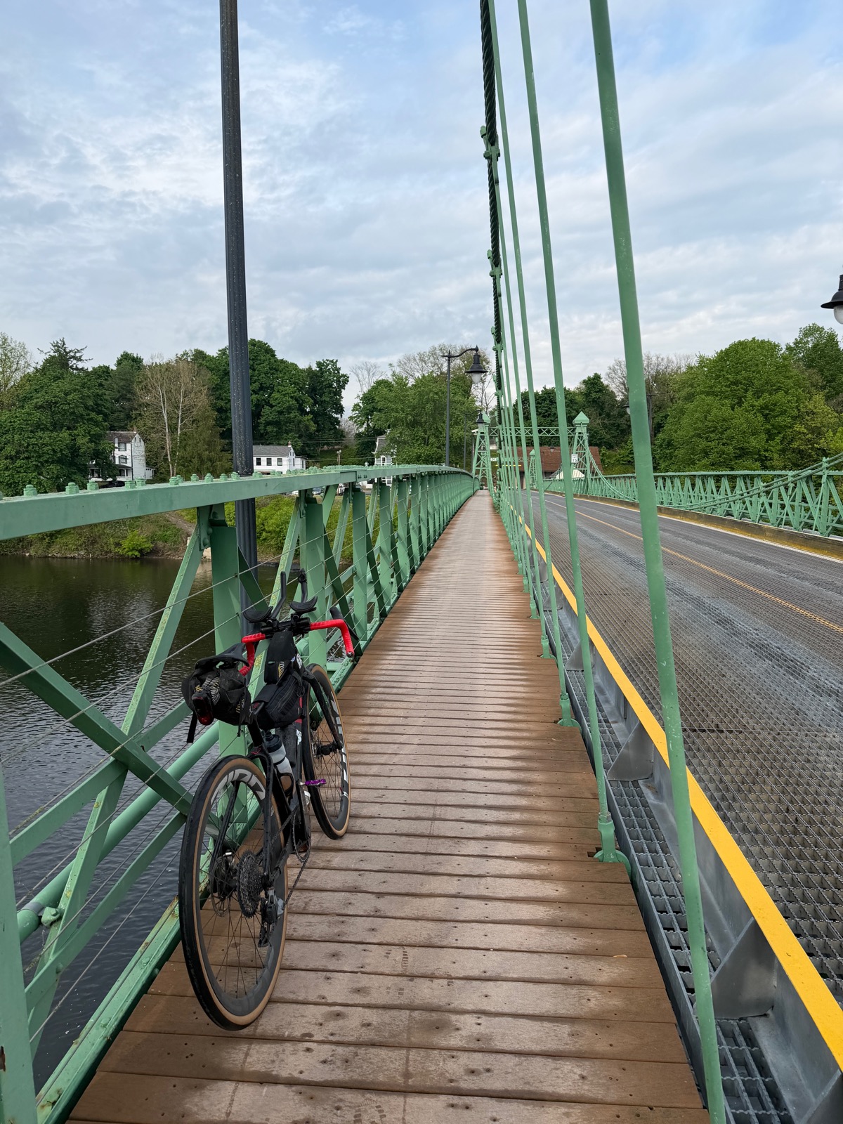 A loaded bike leaning against the railing of a green suspension bridge over the Delaware river, early morning light