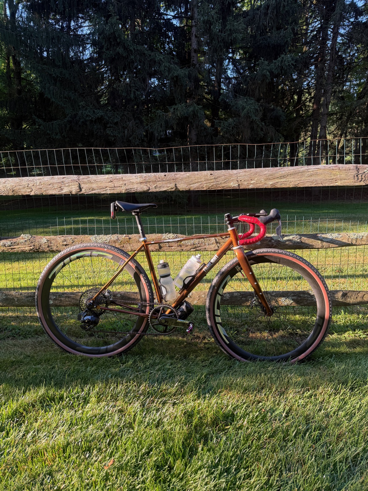 A copper coloured Fairlight Secan with red bar tape and knobby tyres, leaning against a wooden fence in evening light